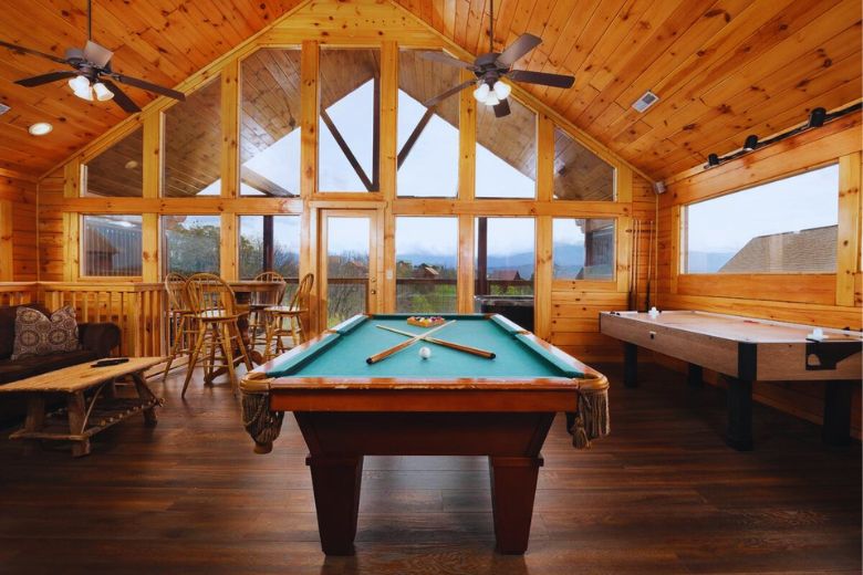 Spacious cabin game room with vaulted wood-paneled ceiling, large floor-to-ceiling windows showcasing a mountain view, a pool table, air hockey table, bar-height table with chairs, and cozy seating area with rustic furniture.